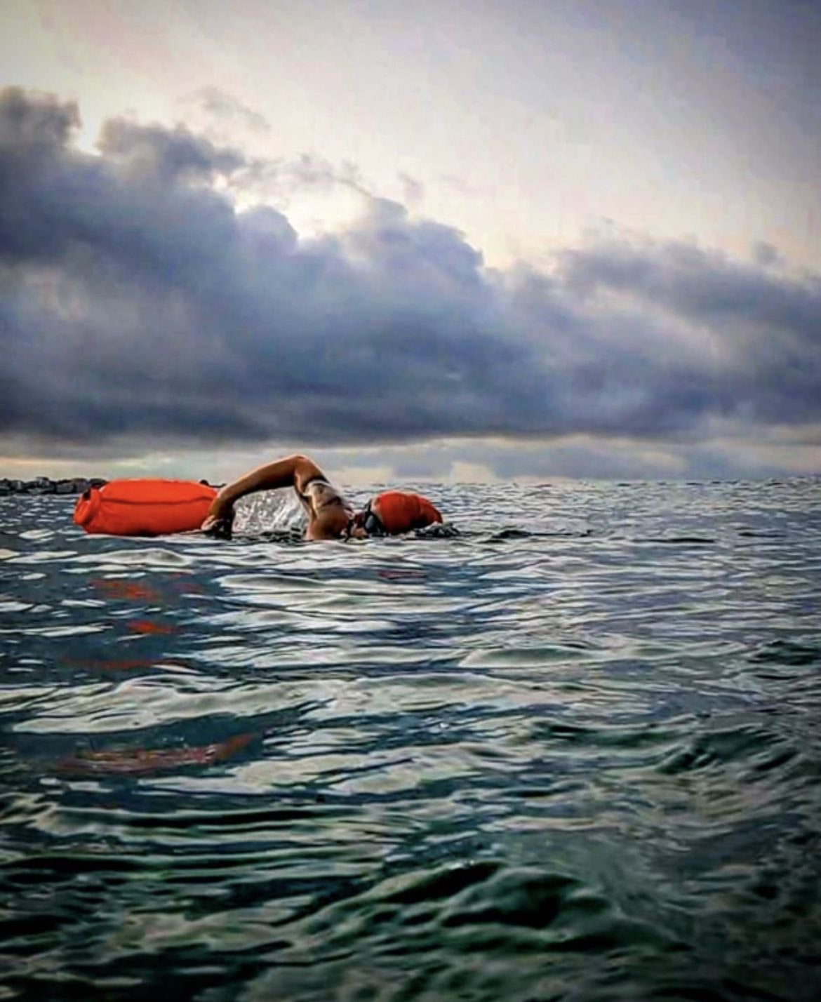 Open-water swimmer with an orange tow float under storm light